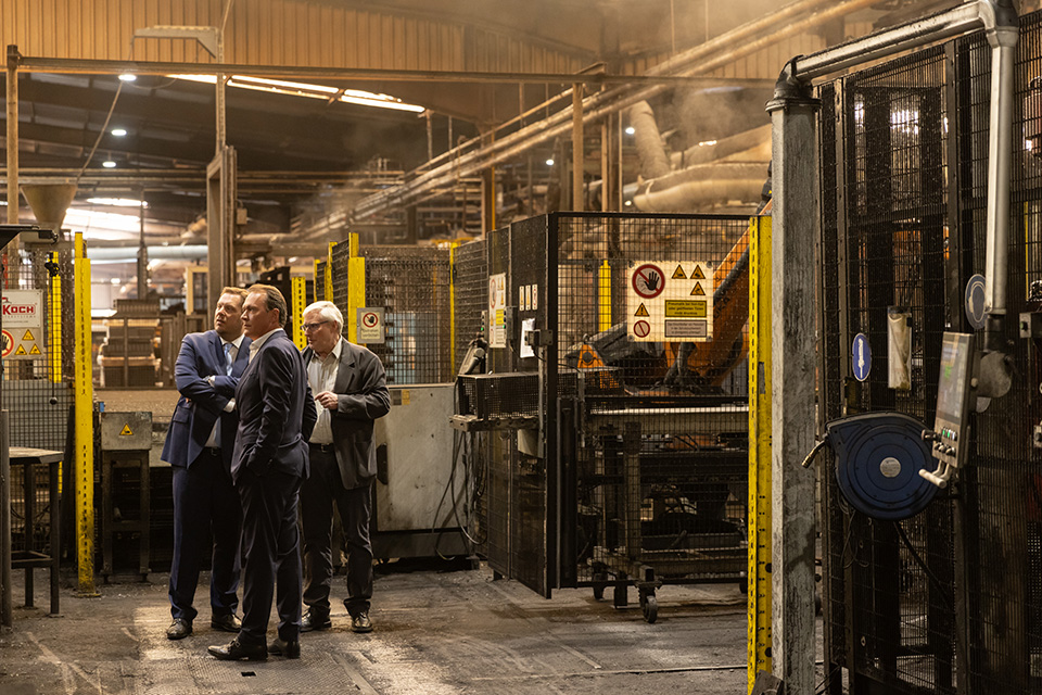 An industrial interior shot shows three standing on a factory floor, observing the machinery around them. From left to right: Daniel Schranz, Mayor of Oberhausen; Matthias Normann, CEO of INTOCAST AG Group; and Manfred Knoll, Plant Director at INTOCAST Oberhausen. They are positioned near yellow and black safety barriers of the presses at the INTOCAST plant in Oberhausen. Steam is visible in the air, creating a soft haze in the background.