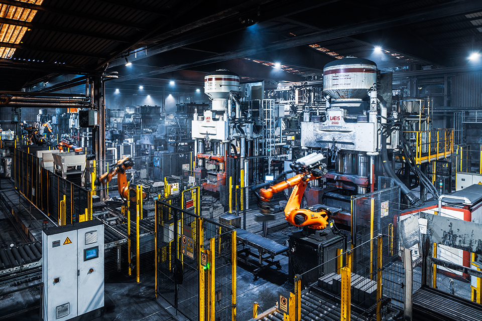 An industrial interior shot of the INTOCAST plant in Oberhausen, Germany, showcasing manufacturing equipment. In the center, two large, LAEIS industrial presses dominate the space. Around them, several bright orange KUKA robotic arms are actively engaged in tasks, positioned on platforms and near conveyor belts behind safety barriers with yellow posts. Overhead, industrial lighting illuminates the scene, and the roof structure of the plant is visible. The overall impression is one of modern, automated heavy industry.