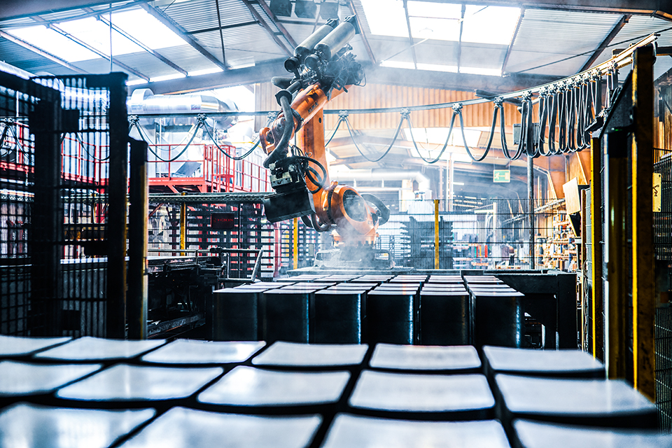 A shot capturing a bright orange KUKA robotic arm in action at the INTOCAST plant in Oberhausen. The KUKA robot arm is shown actively handling dark, rectangular refractory bricks, positioned above a grid of these bricks with steam rising from them, suggesting they’ve just been pressed. In the foreground, a similar grid of finished or prepared rectangular bricks is visible, reflecting light. The background reveals the high-tech, automated industrial setting with metal structures, a conveyor system, and the factory roof with skylights, allowing natural light to filter in, emphasizing a modern production environment for refractory materials.